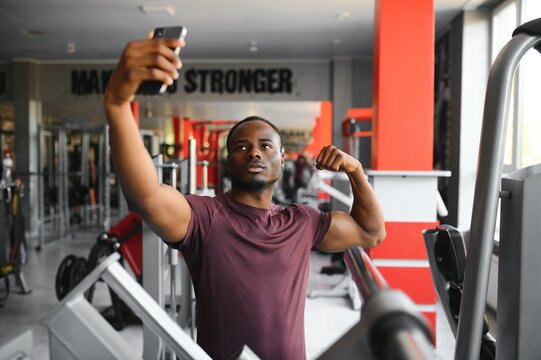 smiling young sportsman showing biceps and taking selfie with smartphone in gym.