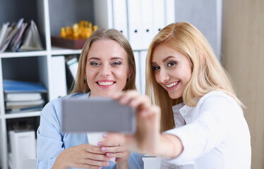 Two businesswomen in the office smile and do selfie on a smartphone during a break to have coffee