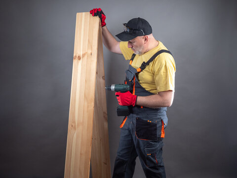 Furniture assembler with boards. Man uses electric screwdriver. Worker in gray suit. Builder with screwdriver. Construction company worker. Man carpenter on black. Master with parquet board
