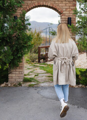 Fototapeta na wymiar Back view, lifestyle stylish young woman with blonde hair woman in beige oversized trench coat, blue jeans and white boots walking around the city on an autumn sunny day, autumn lifestyle
