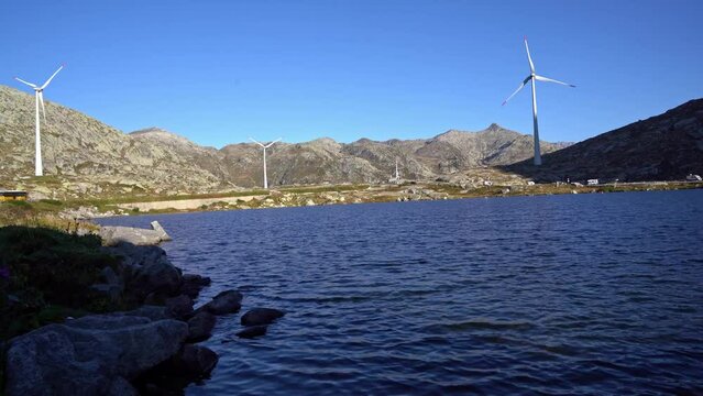 Mountain lake with wind turbines in the background at summit of Swiss mountain pass Gotthard on a sunny late summer morning. Movie shot September 10th, 2023, Gotthard, Switzerland.