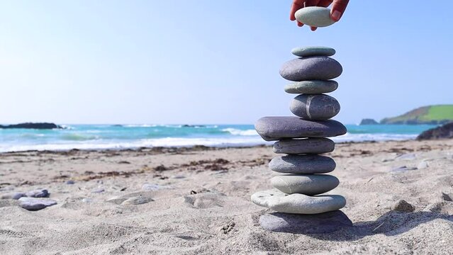 Zen stack of stones on a beach showing balance. A hand tries to place another stone but the stack collapses.