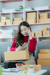 Asian woman small business owner starting an SME. Independent Asian woman using laptop working with boxes at home. Young Asian woman who succeeds by raising her hand Packaging boxes, online marketing 