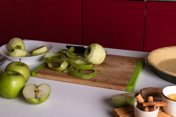 One green pleeling apple on a cutting board and kitchen knife on a table with ingredients for apple pie: green apples, cinnamon sticks and raw pie dough. Thanksgiving preparation,