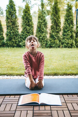 Fototapeta premium Little girl sitting in Simhasana or Lion Pose on the roll mat practicing yoga outdoors