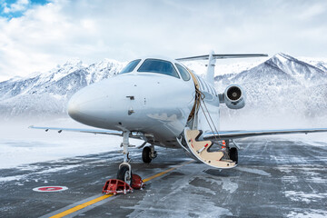 Fototapeta na wymiar Modern white executive aircraft with an opened gangway door at the winter airport apron on the background of high scenic snow capped mountains