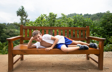 A happy child in the summer heat lies on a wooden bench against the backdrop of tropical plants. The boy is on vacation in the summer.