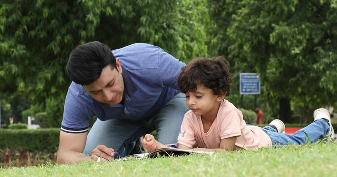 Video of father talking to his cute son while lying on grass in garden drawing pictures with him.
