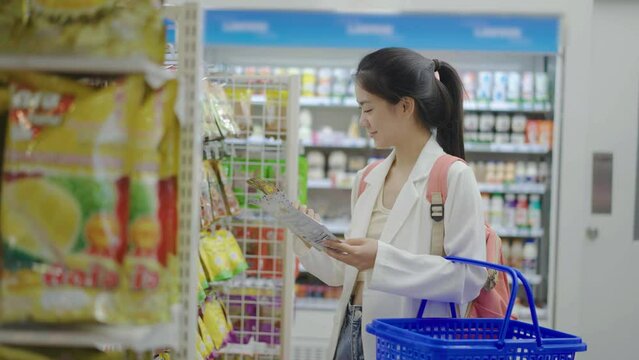 Young asian woman buy snacks and dried fruits at store, She stand against shelves and look to product details. Then search for required goods on racks, pick it up and put into shopping cart