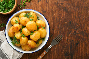 Boiled new or young potatoes with butter, fresh dill and onions in white bowl on dark old rustic wooden background. Tasty new boiled potatoes. Top view.