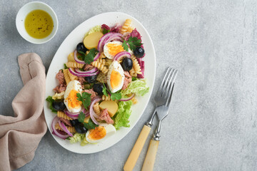 Tuna salad with pasta, eggs, potatoes, olives, red onions and sauce in white plate on old light gray concrete table background. Nicoise salad. French cuisine. Top view. Flat lay.