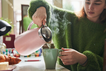 Calm teen girl pouring hot tea while sitting at kitchen during breakfast - 646385853