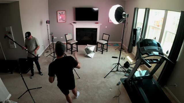 Two caucasians videographers setting up camera equipment indoor for a two person interview. Working with tripods, cameras, lights, boom mics, lavalier microphones, sand bags and chairs for guests.
