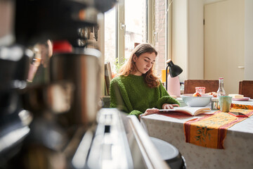 Young teen woman enjoying exciting novel while having hot drink and tasty breakfast in kitchen