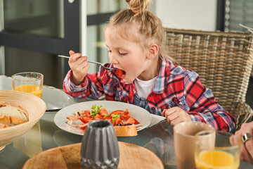 Focused child girl holding fork and eating with appetite while enjoying healthy breakfast