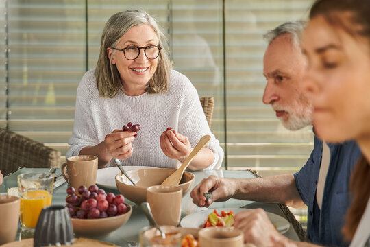 Cheerful grandmother in eyeglasses holding grape and looking at her family