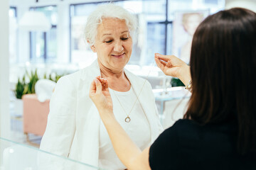Fototapeta premium Sale, consumerism, shopping and people concept. Grey haired elderly woman buying some jewelry. Woman owner, saleswoman of jewelry shop presenting for senior female consumer golden chain with pendant.