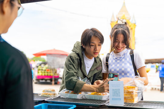 Group of Happy Asian people tourist buying and eating street food with using mobile app on smartphone scan QR code making online payment during travel local town in Thailand on summer holiday vacation - Powered by Adobe