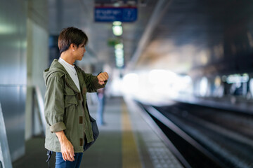 Young Asian man using smart watch during waiting for train at railway station. Handsome guy enjoy outdoor lifestyle travel in the city with using wireless technology on summer holiday vacation.