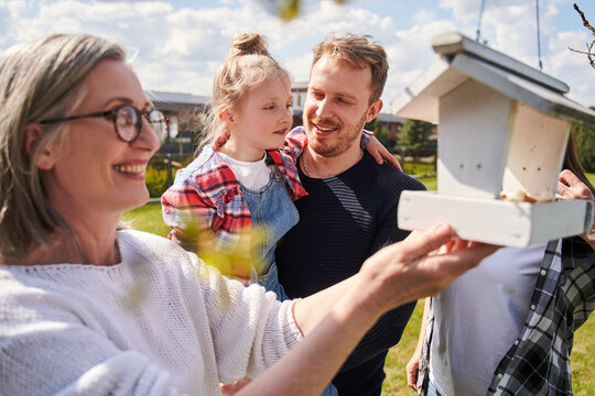 Cheerful grandmother putting food at wooden birdhouse while her son holding little girl