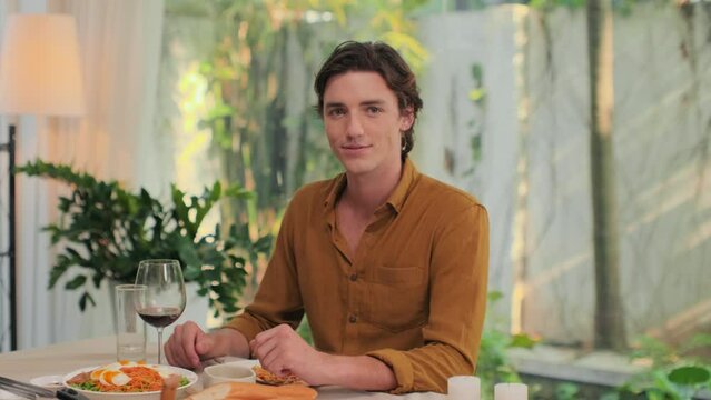 Portrait of young Caucasian man sitting at dinner table, drinking red wine and looking at camera