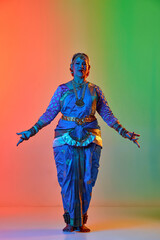 Fototapeta premium Beautiful indian woman dancing traditional dance in elegant blue dress against gradient studio background in neon light. Concept of beauty, fashion, India, traditions, choreography, art. Ad