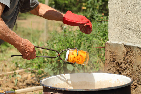 Man hands, one with red glove, holding an old canning jar lifter, drawing a jar with apricot slices out of boiling water in a big rustic pot. Bath canning fruits outside.