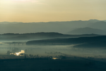 Aerial view of the valley in early morning mist, beautiful in the highlands. Low clouds and fog cover the sleeping meadow. Alpine mountain valley mists landscape at dawn. Serene moment in rural area