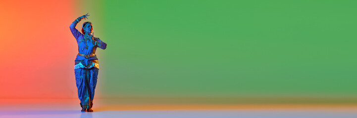 Mature woman dancing traditional dance in elegant blue dress over gradient studio background in neon light. Concept of beauty, fashion, India, traditions, choreography, art. Copy space for ad. Banner