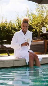 Vertical video portrait of woman wearing robes outdoors sitting and relaxing with glass of champagne by swimming pool with feet in water on spa day - shot in slow motion