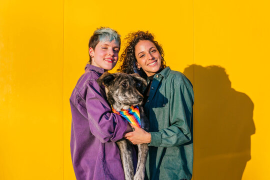 Smiling lesbian couple carrying dog in front of yellow wall
