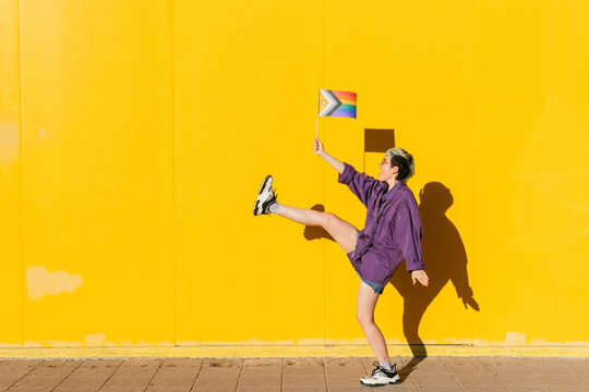 Lesbian woman dancing with one leg holding rainbow flag in front of yellow wall