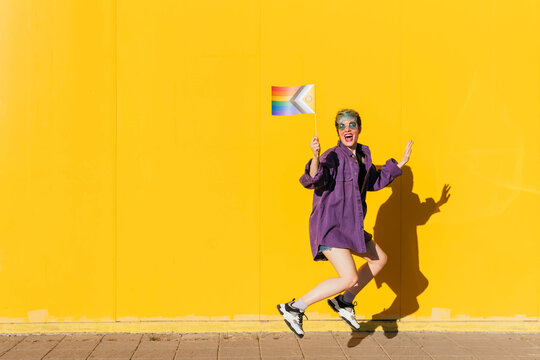 Cheerful lesbian woman with rainbow flag enjoying in front of yellow wall