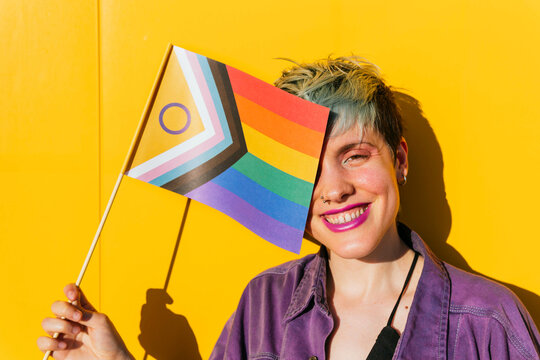 Cheerful lesbian woman covering eye with rainbow flag in front of yellow wall