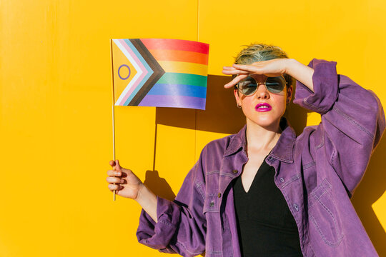 Lesbian woman shielding eyes and holding rainbow flag in front of yellow wall