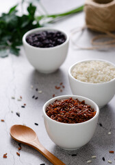 Red, black, and Yamani rice in white bowls, a wooden spoon and a rustic thread, on grey stony background.