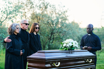 Young pastor with Holy Bible and family of three standing by covered coffin with white flowers on...