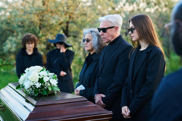 Family of three grieving people in sunglasses and mourning clothes standing in front of coffin with...