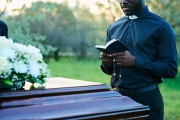 Cropped shot of young priest in black clothes reading verses from Holy Bible during funeral service...