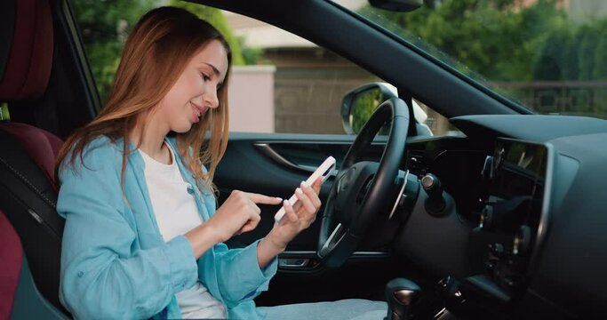 Young woman is sitting in the driver's seat using a touch display screen multimedia device in sporty car driven. Young girl sitting behind wheel of premium modern car with smartphone in her hands.