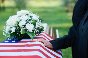 Hand of mature woman in mourning attire on American flag covering coffin with bunch of resh white...