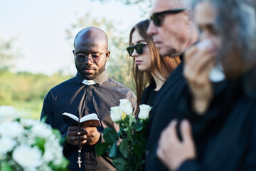 Focus on serious priest in black attire looking in open Holy Bible at funeral service while reading...