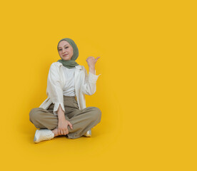 Young caucasian muslim woman in hijab sit floor legs crossed full body view point thumb finger aside copy space workspace area. Isolated on plain yellow background studio portrait. Advertising concept