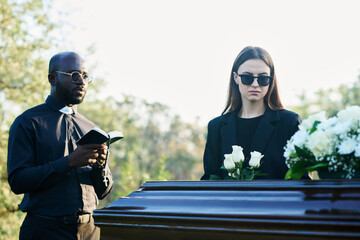 Young inconsolable widow in mourning attire and sunglasses holding bunch of white roses while...