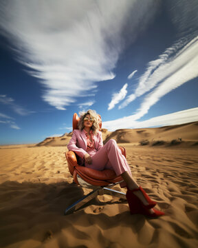 An elegant blonde woman wearing all pink suit and sunglasses, sitting in armchair in the middle of the desert. Surreal concept for adventure or holidays in the desert. Ad for travel agency. Generative