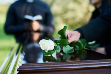Fresh white rose being put by woman in mourning attire on top of closed coffin lid against priest with open Bible carrying out funeral service - obrazy, fototapety, plakaty