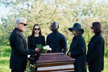 Mature man in black suit and sunglasses putting bunch of white roses on top of closed lid of coffin...