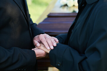 Close-up of hands of mature grieving man on those of his mourning wife in black attire standing in...