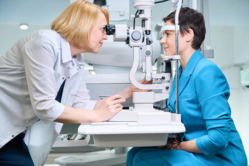 Experienced oculist checking adult woman vision with ophthalmic equipment - obrazy, fototapety, plakaty