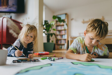 Siblings lying on stomach and painting at home with watercolors, markers and tempera paints,...